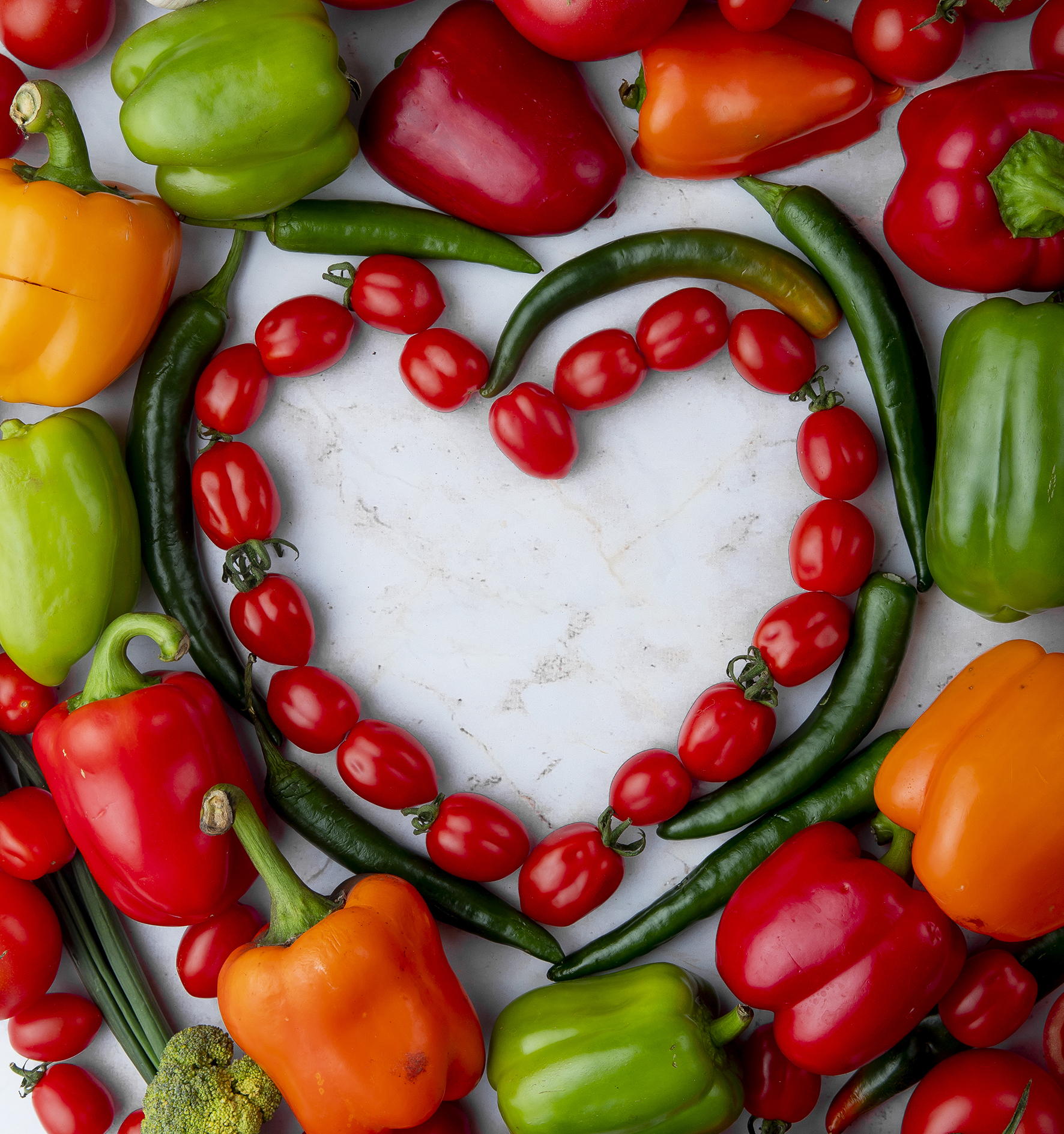top view of fresh ripe vegetables arranged in a heart shape cherry tomatoes green chili pepper bell pepper garlic and onion on marble background
